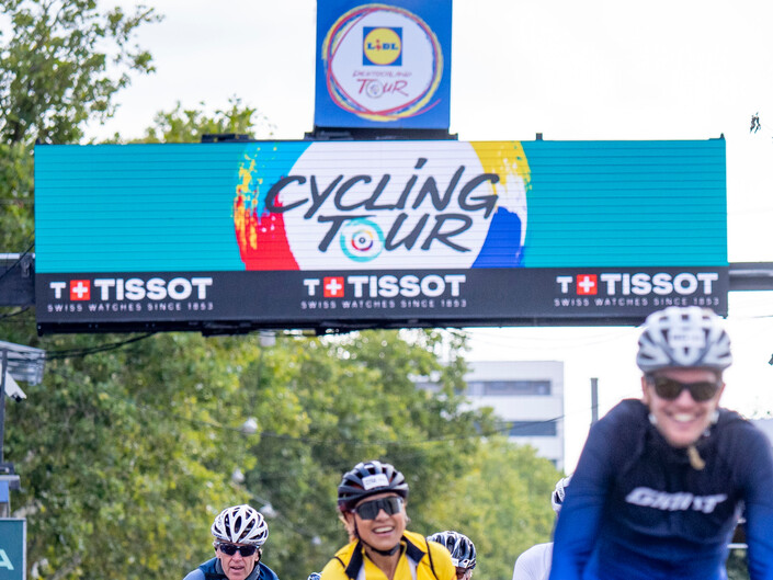 Radler fahren auf den Betrachter zu im Hintergrund ist das Logo der Cycling Tour zu sehen