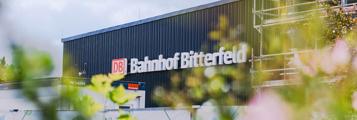 Eine Blumenwiese im Vordergrund verdeckt Teile des Bahnhofsgebäudes