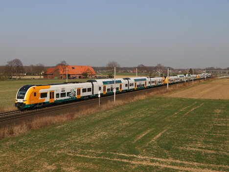 Ein weiß-grün-orangener Doppelstockzug vom Typ Desiro HC fährt durch eine offene Landschaft mit Feldern und einigen Häuser.n