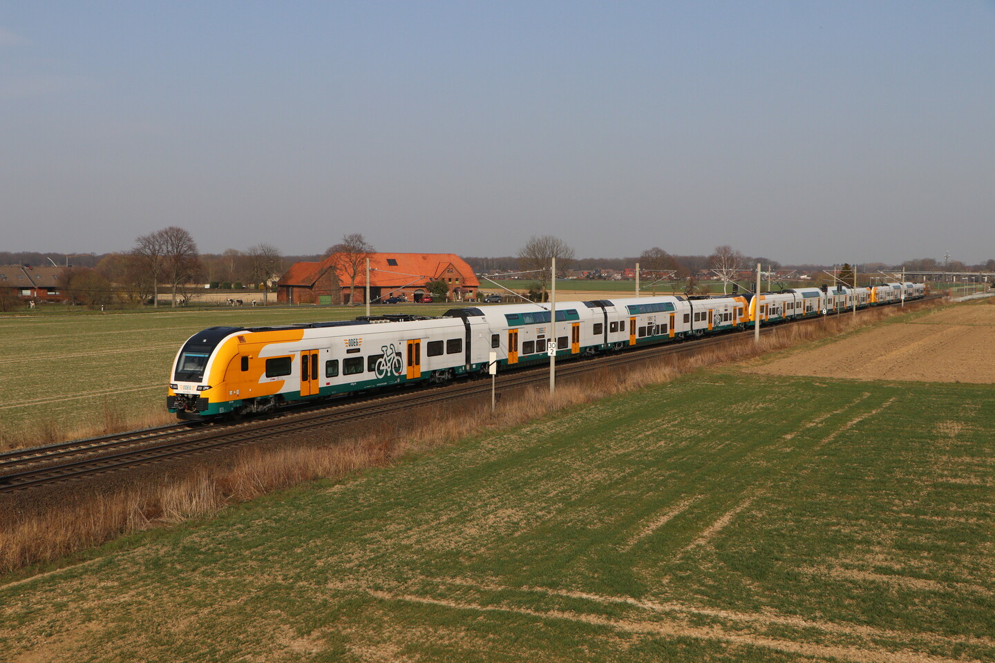 Ein weiß-grün-orangener Doppelstockzug vom Typ Desiro HC fährt durch eine offene Landschaft mit Feldern und einigen Häuser.n