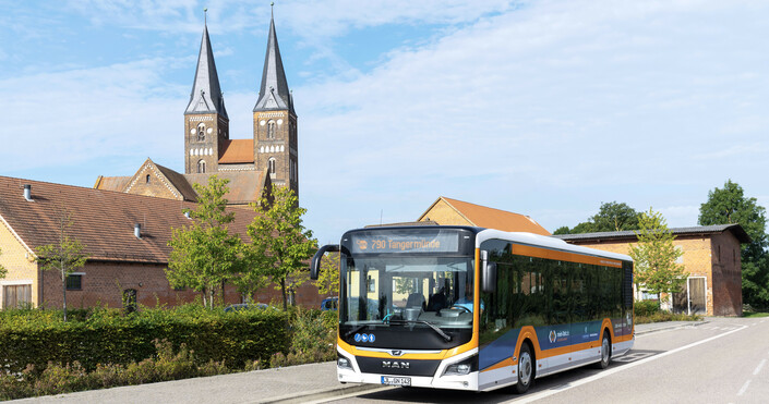 Ein orange-schwarzer PlusBus fährt durch Jerichow. Im Hintergrund ist das Backsteinkloster zu sehen